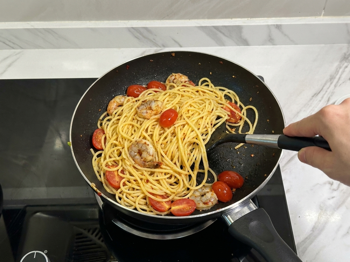 Una spatola nera che salta spaghetti, gamberetti conditi e pomodorini tagliati a metà in una padella.