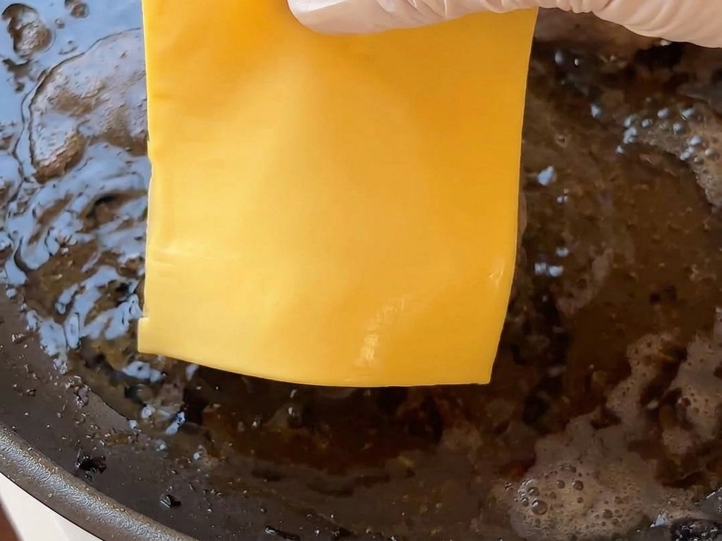 Una mano che posiziona una fetta di formaggio giallo sopra un burger bollente in padella.