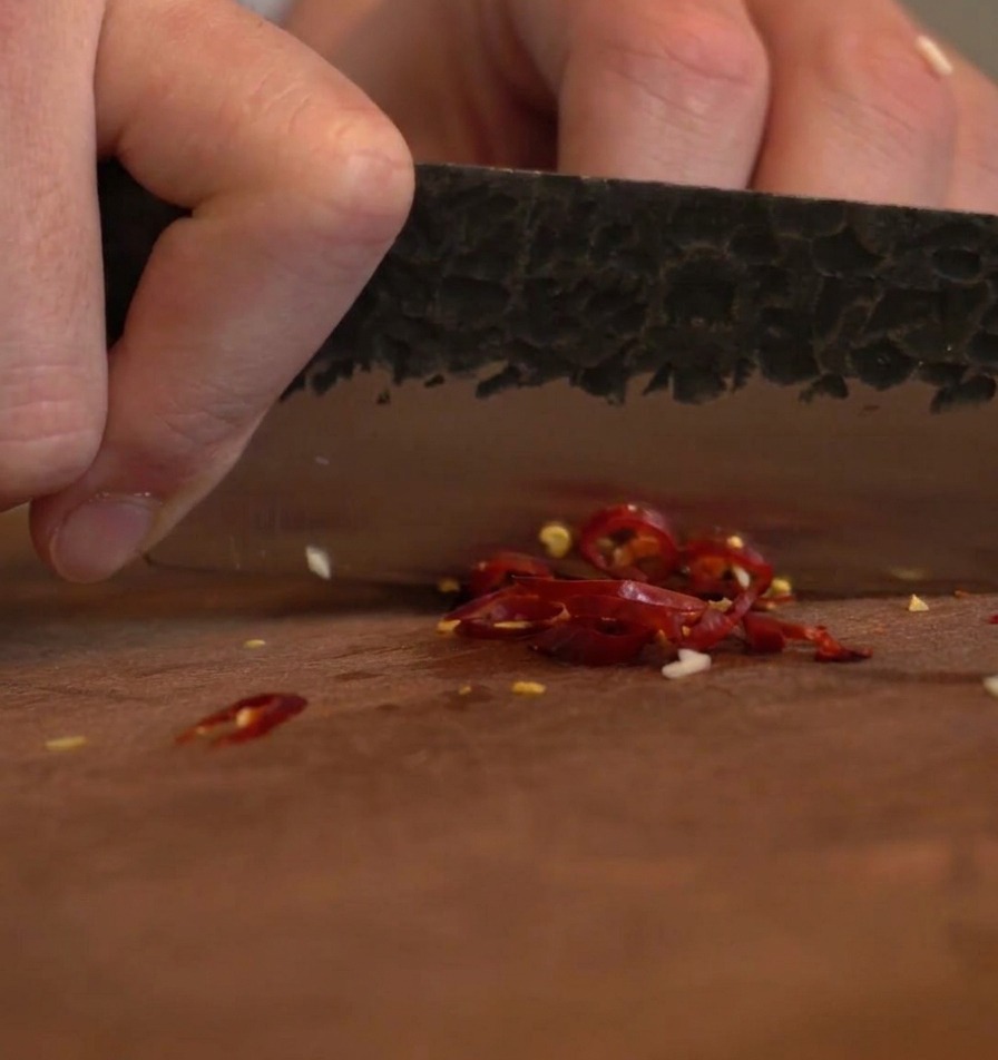 La lama scura e ruvida di un coltello che trita vivaci peperoncini rossi secchi su un tagliere di legno.
