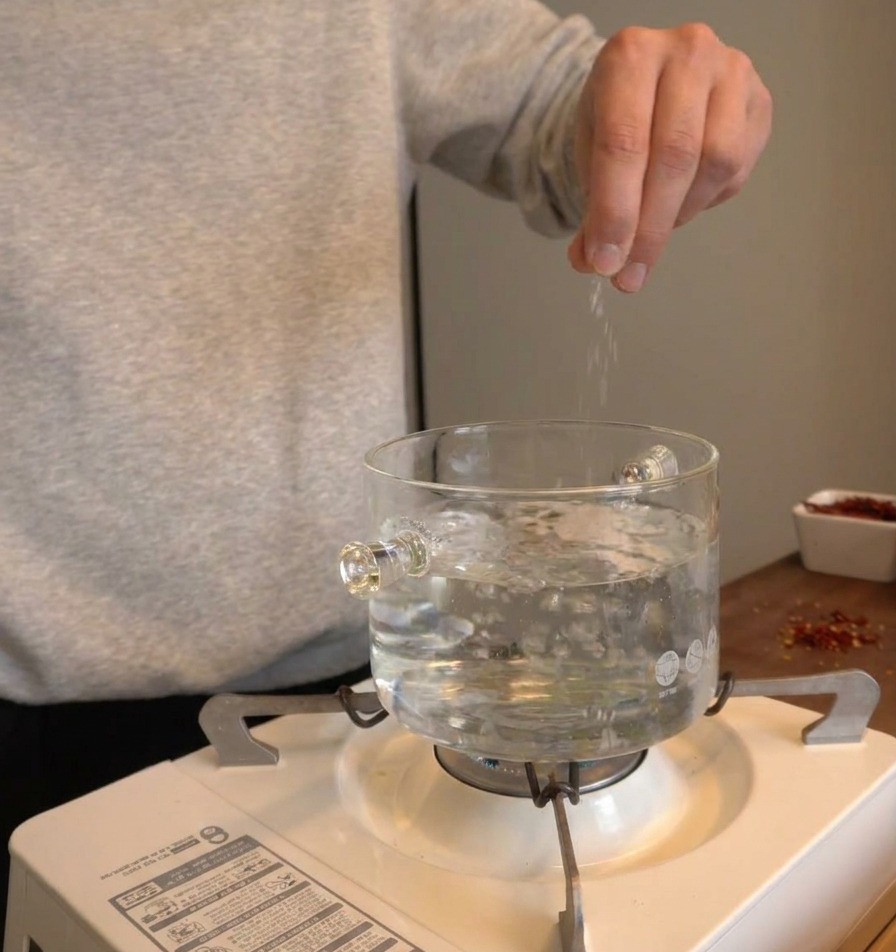 Mano che sparge sale in una pentola di vetro trasparente con acqua in piena ebollizione su un fornello portatile.