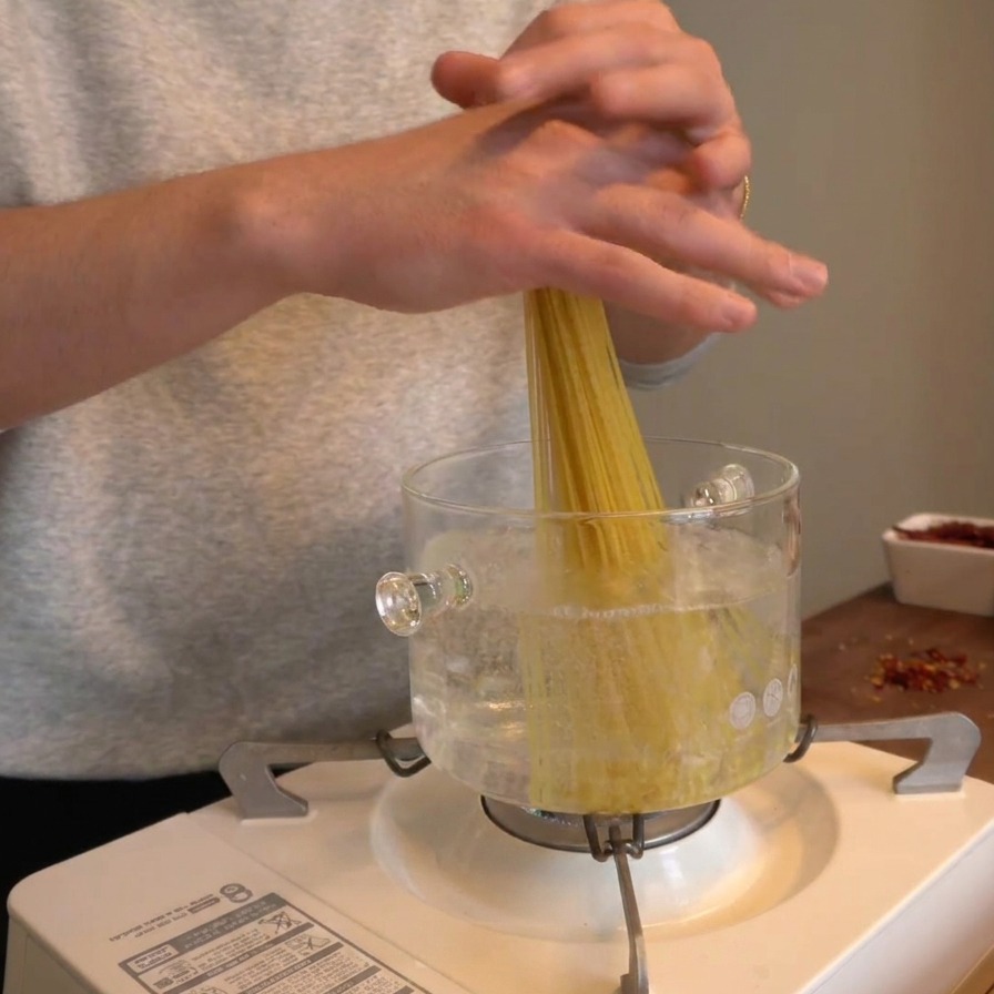 Mani che fanno scivolare verticalmente un mazzetto di spaghetti secchi in una pentola di vetro con acqua bollente.