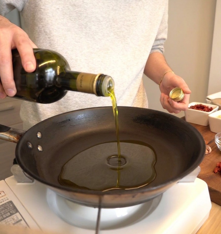 Olio doliva versato da una bottiglia verde scuro in una padella nera.