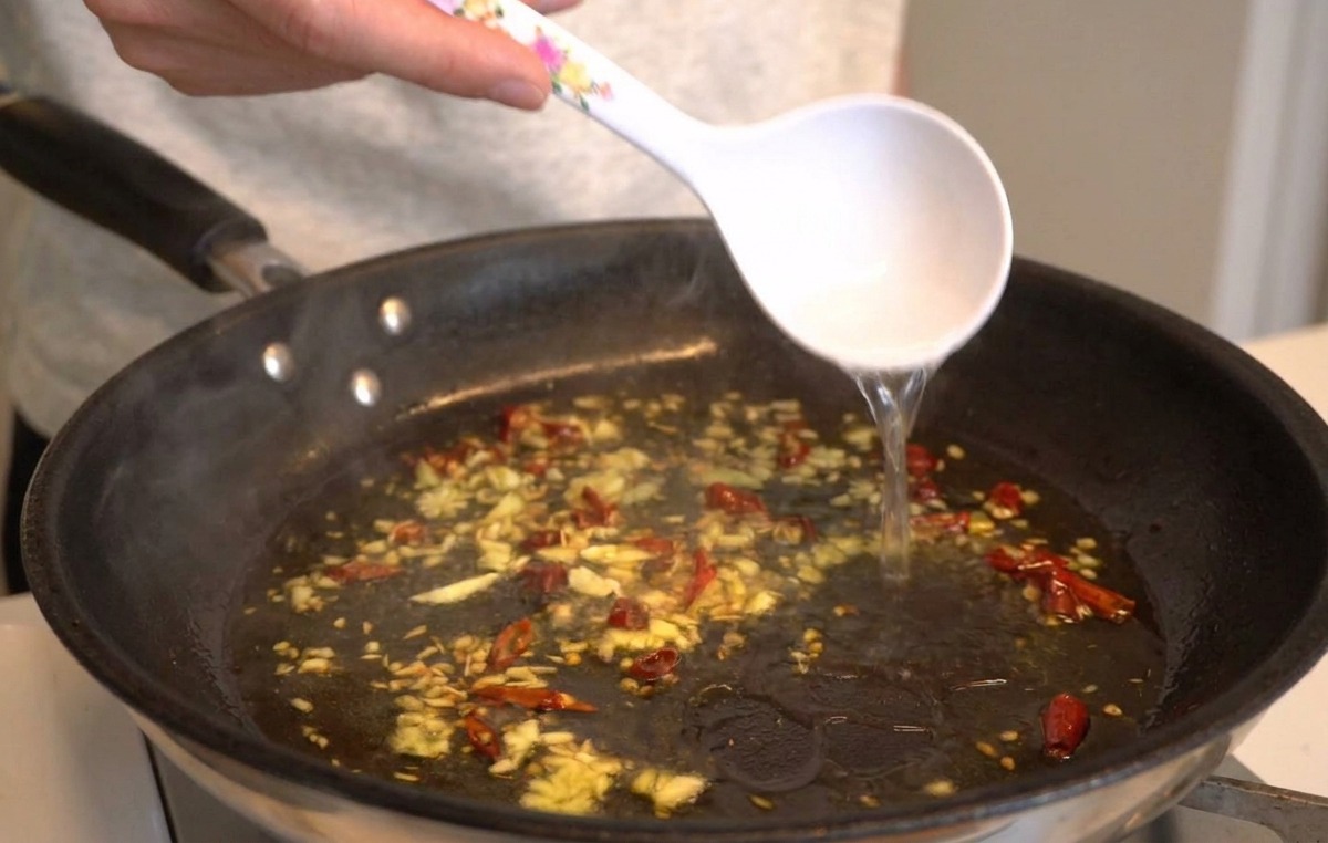 Un mestolo bianco che versa acqua calda e ricca di amido in una padella contenente olio, aglio sfrigolante e peperoncini.