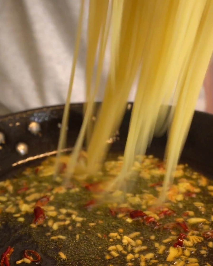 Lunghi fili di spaghetti trasferiti in una padella in cui sobbolle il condimento di olio, aglio e peperoncino.