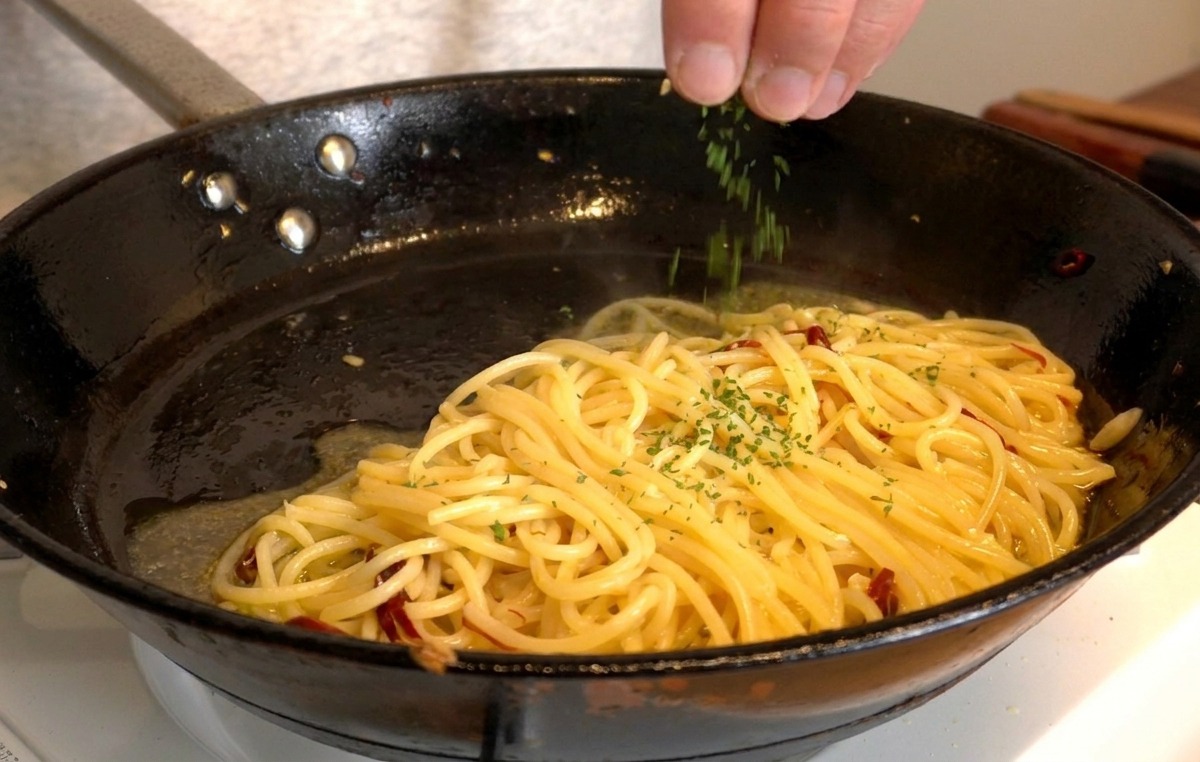 Una mano che cosparge del prezzemolo verde appena tritato su una padella di spaghetti lucidi.