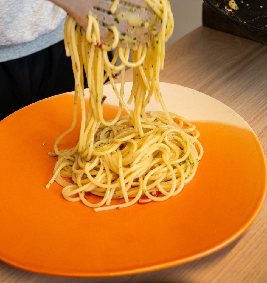 Pinze che sollevano e arrotolano gli spaghetti aglio e olio su un piatto da portata arancione brillante.
