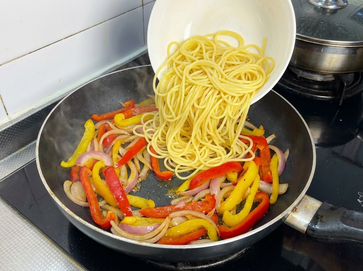 Spaghetti bolliti versati sopra peperoni e cipolle in padella.