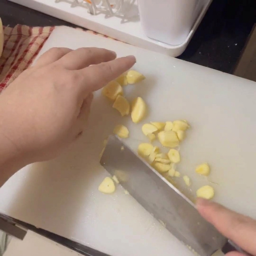 Mani che usano un coltello per affettare finemente spicchi daglio fresco su un tagliere bianco.