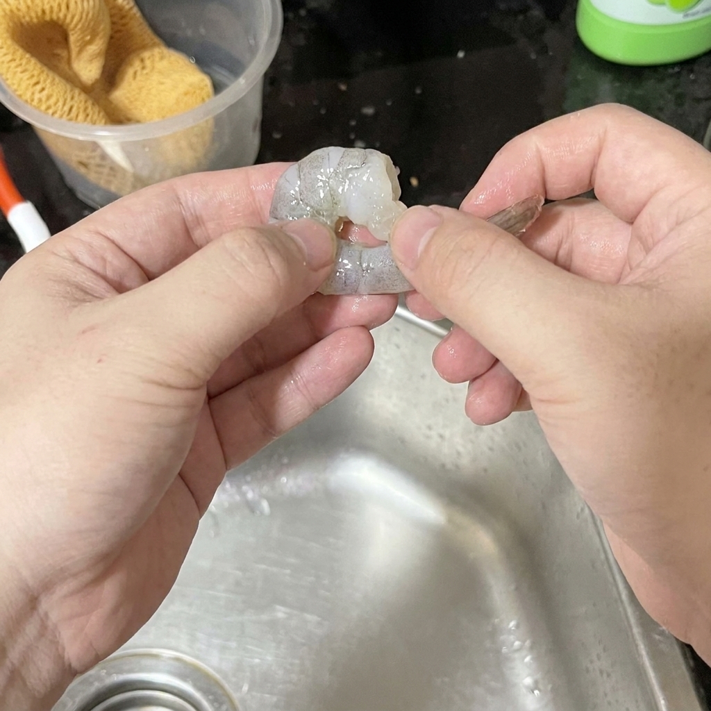 Due mani che sbucciano con cura un gambero grigio crudo sopra un lavandino in acciaio inox.