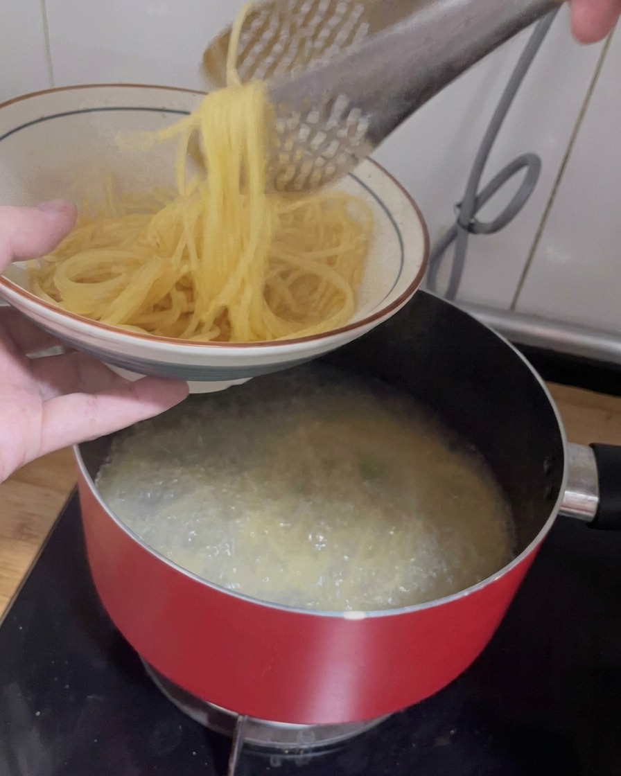 Pinze da cucina che sollevano spaghetti cotti da una pentola verso una ciotola in ceramica.