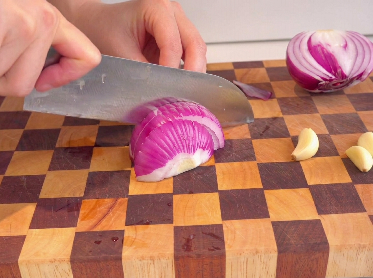 Mani che usano un coltello per tritare una cipolla rossa su un tagliere a scacchi.
