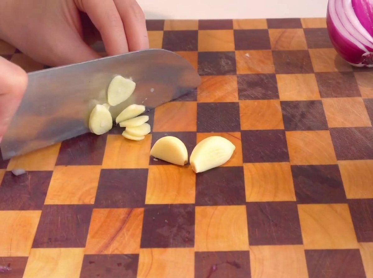 Mani che affettano spicchi daglio sbucciati su un tagliere di legno a scacchi.