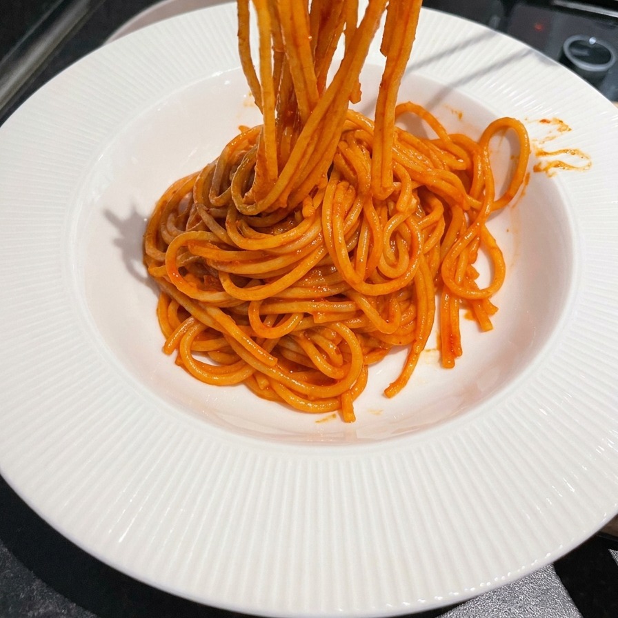 Bacchette di metallo che sollevano e arrotolano spaghetti rossi conditi in una ciotola bianca.
