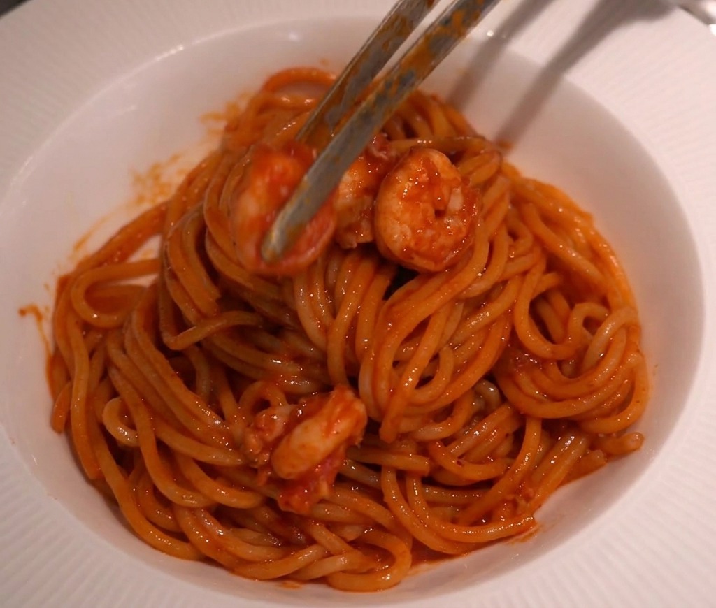 Pinze da cucina che adagiano i gamberi sopra un nido di spaghetti in una ciotola bianca.