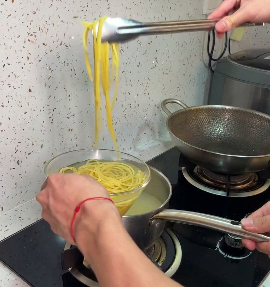 Pinze da cucina che trasferiscono gli spaghetti cotti dalla pentola a una ciotola di vetro con acqua fredda.