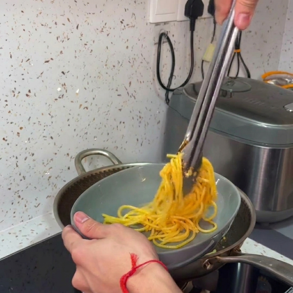 Mani che usano pinze per sollevare e arrotolare gli spaghetti conditi in una ciotola grigia.