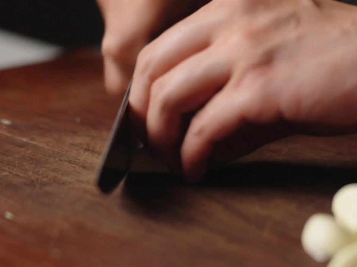 Un coltello che affetta spicchi daglio sbucciati su un tagliere di legno scuro.