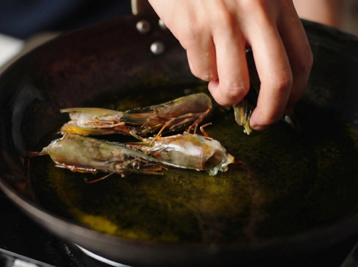 Una mano che mette teste di gamberi crudi in un wok pieno di olio bollente.
