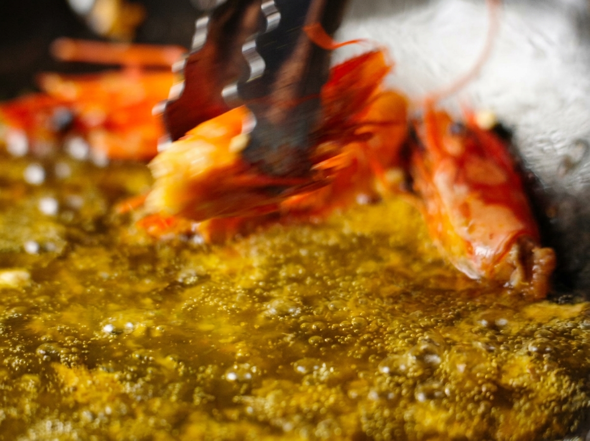 Pinze che sollevano una testa di gambero fritta da una padella di olio infuso color arancio.