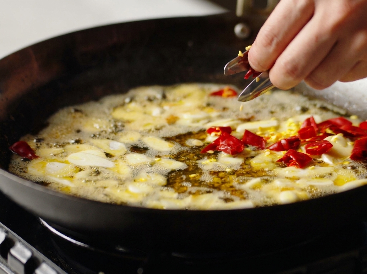 Una mano che aggiunge pezzi di peperoncino rosso secco in un wok con aglio e olio.