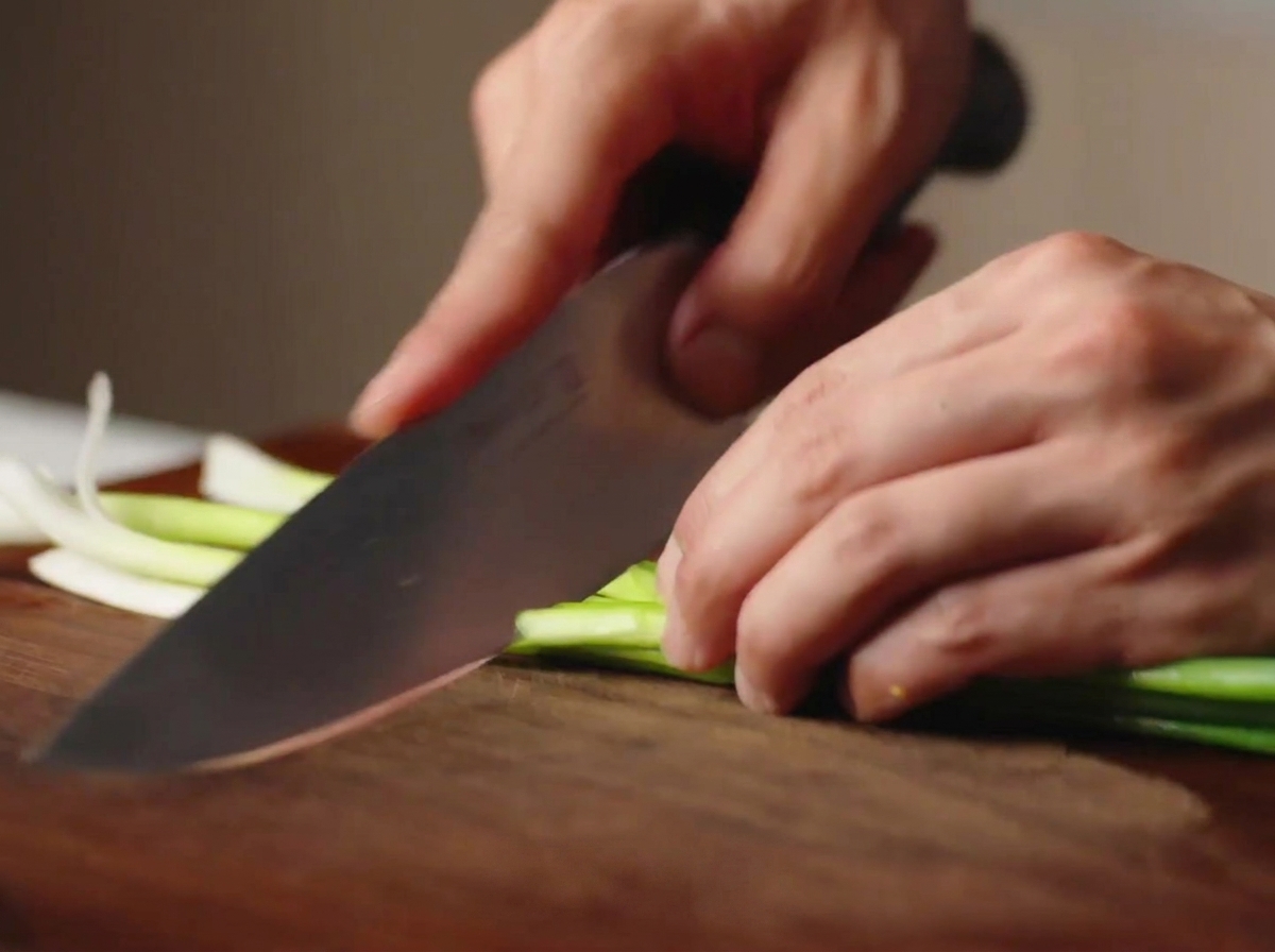 Mani che usano un coltello da chef per tagliare cipollotti freschi a segmenti su un tagliere di legno.