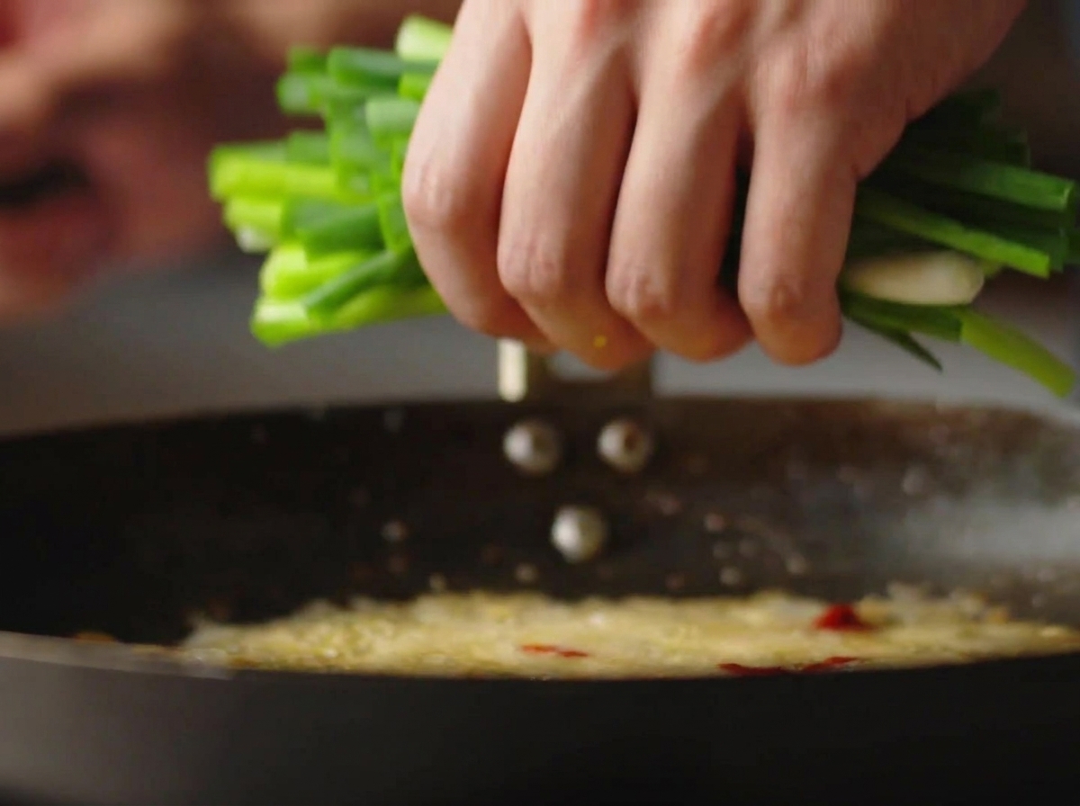 Una mano che versa una manciata di cipollotti freschi in un wok con olio bollente.