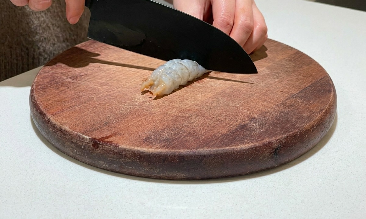 Un coltello che incide il dorso di un gambero crudo sgusciato su un tagliere di legno.