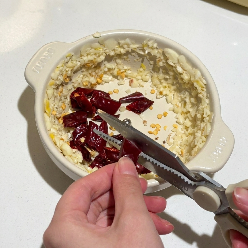 Mani che usano forbici da cucina per tagliare peperoncini rossi secchi in una ciotolina con aglio tritato.