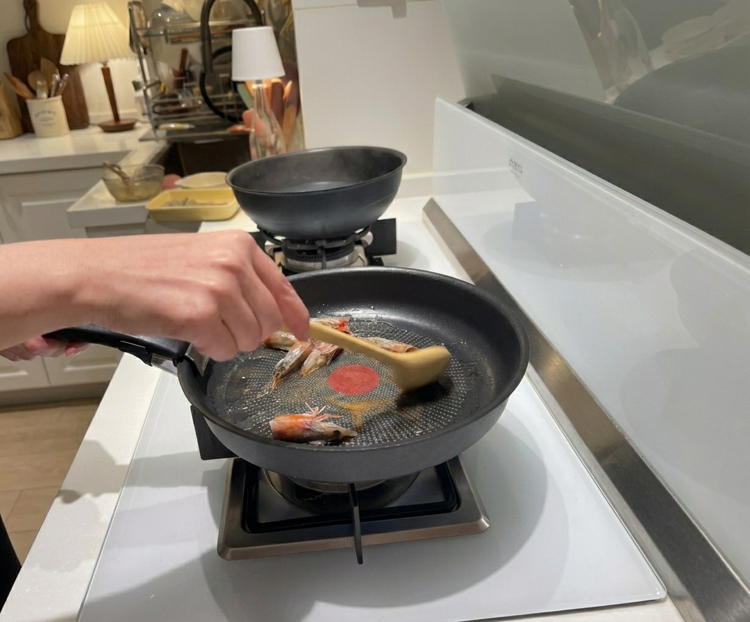 Una mano che usa un cucchiaio di legno per soffriggere le teste di gambero crude in olio caldo dentro una padella nera.