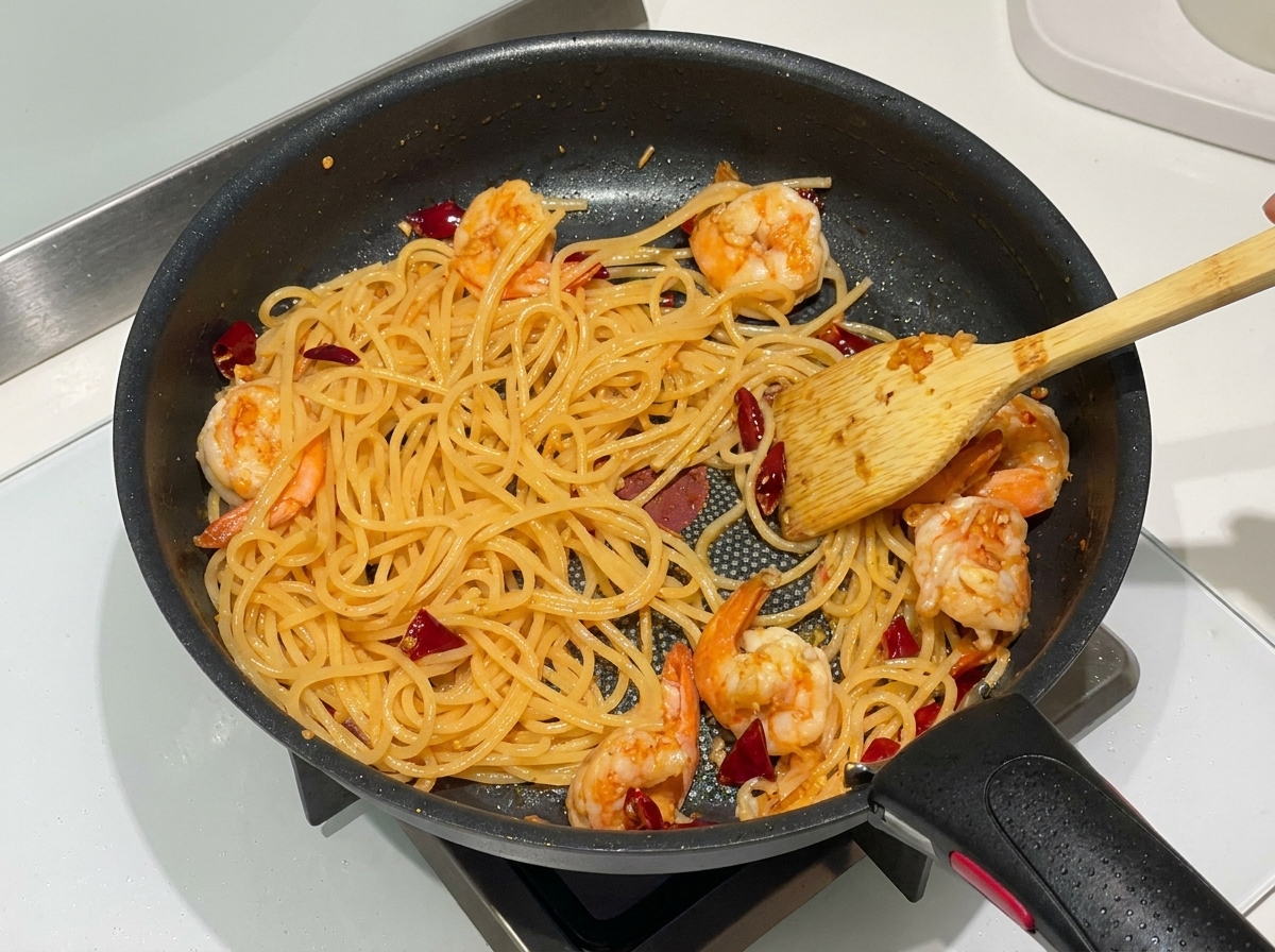 Spaghetti cotti, gamberi rosa arricciati e peperoncini secchi saltati insieme in una padella antiaderente nera con una spatola di legno.