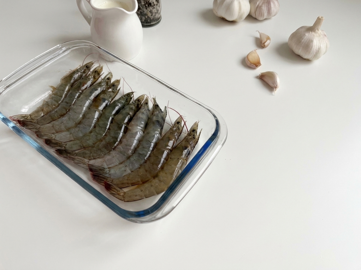 Gamberi grigi crudi e traslucidi disposti in una pirofila di vetro.