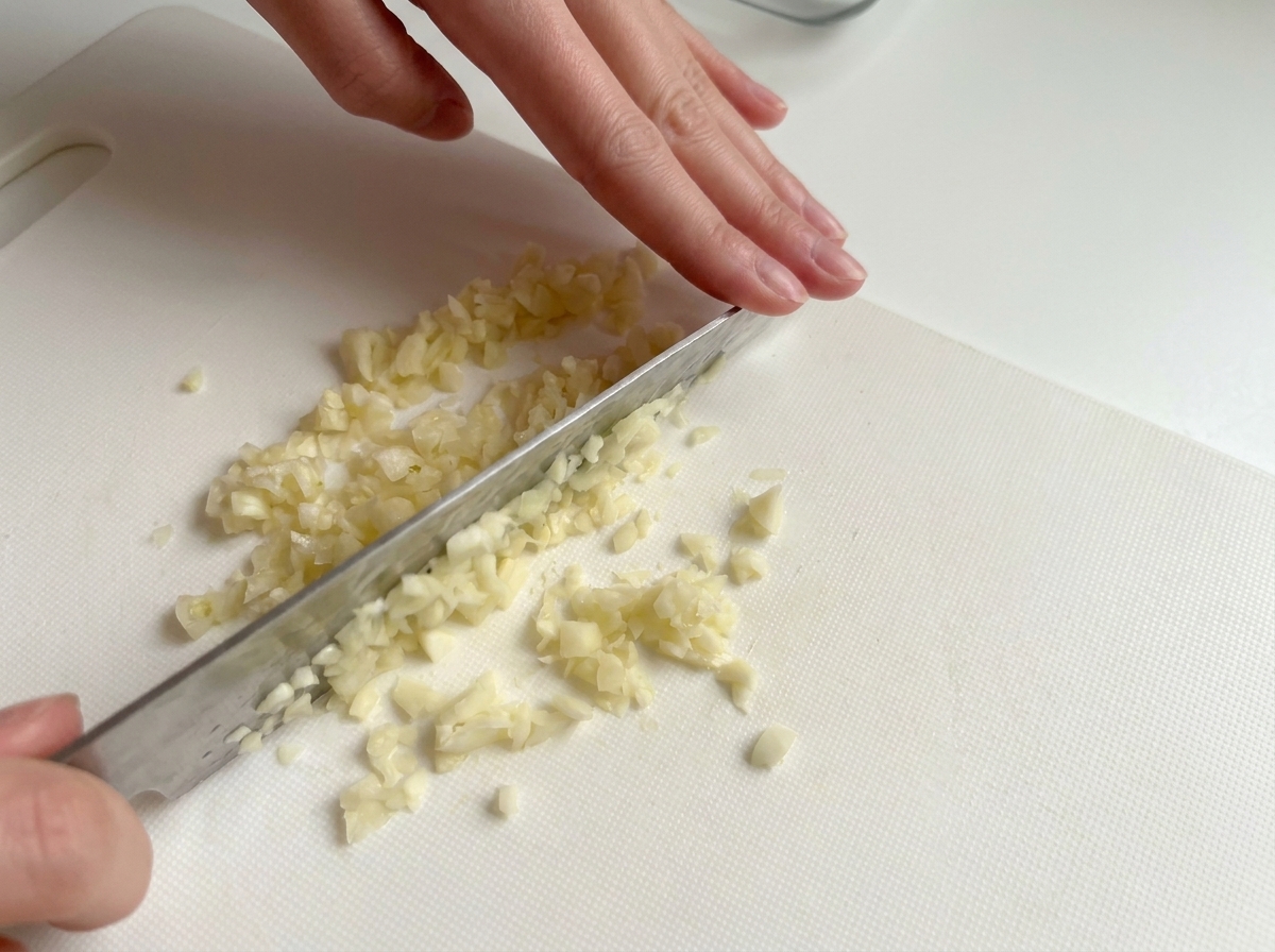 Mani che tritano finemente spicchi daglio su un tagliere bianco.