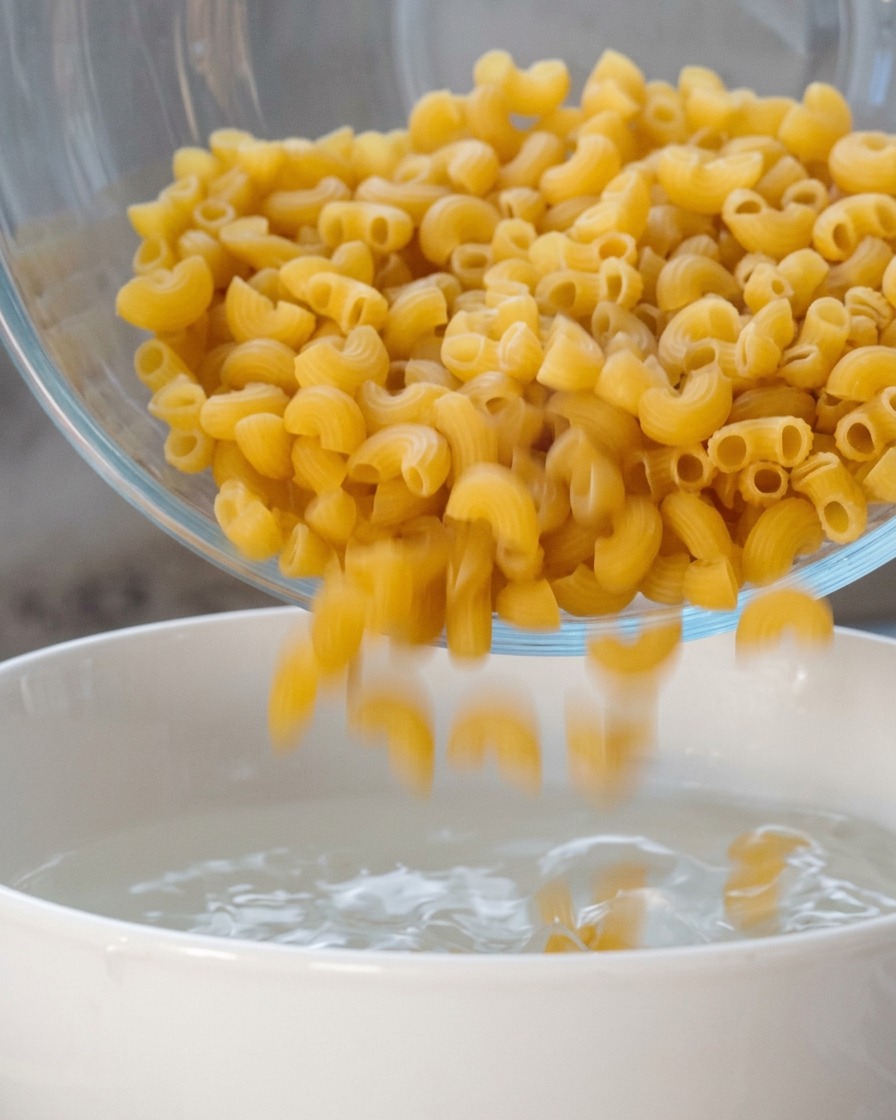 Fusilli secchi versati da una ciotola di vetro in una pentola bianca con acqua bollente.