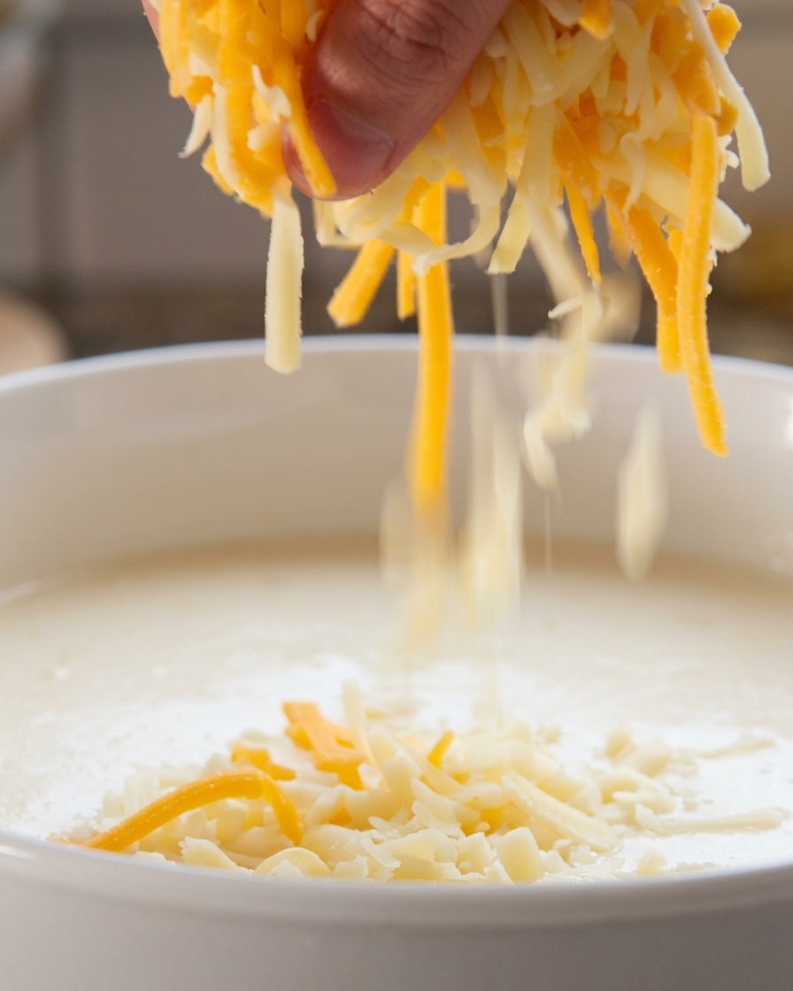 Una mano cosparge un mix di formaggio grattugiato bianco e arancione in un liquido bianco che sobbolle.