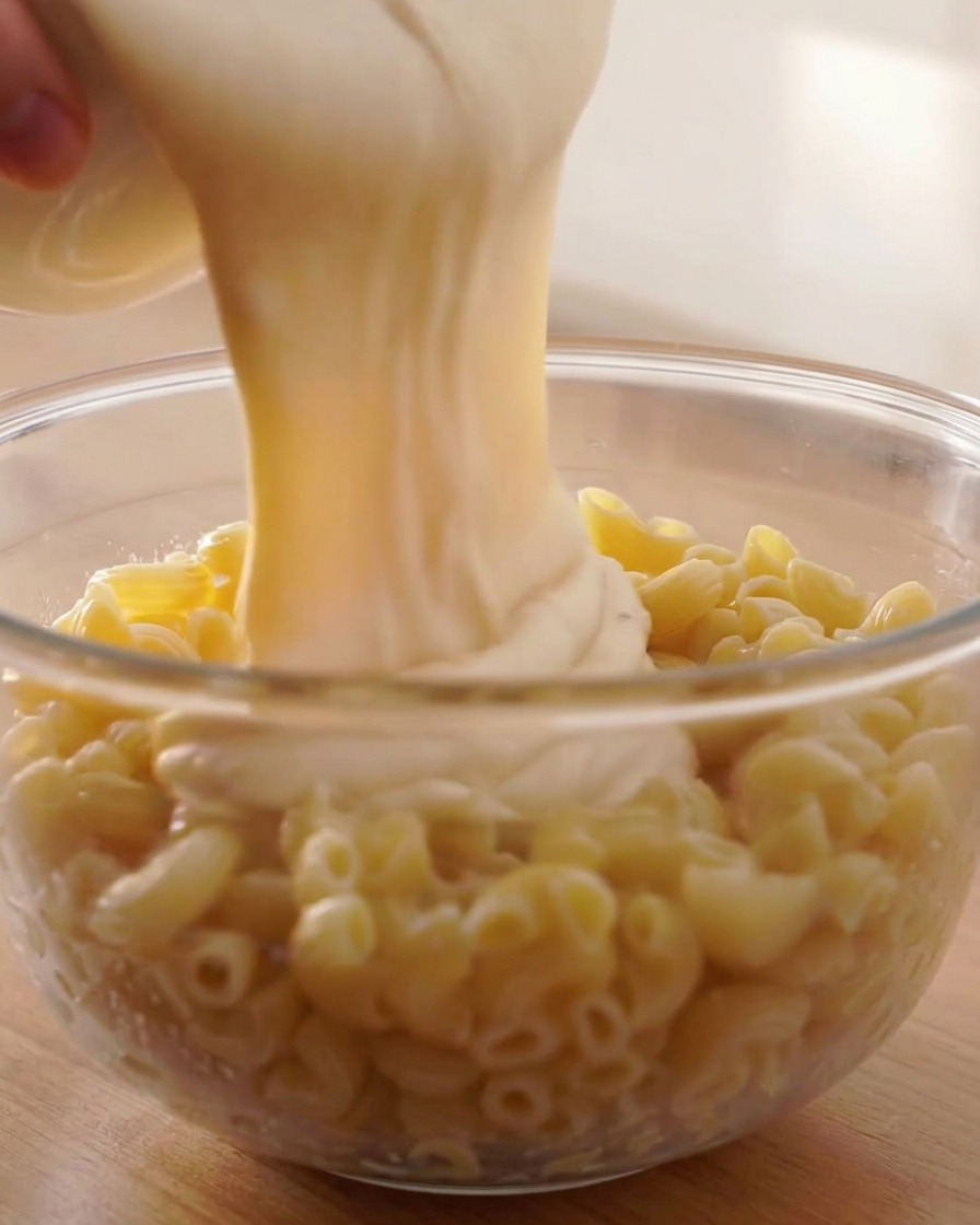 Densa salsa al formaggio versata da un contenitore bianco sopra fusilli cotti in una ciotola di vetro.