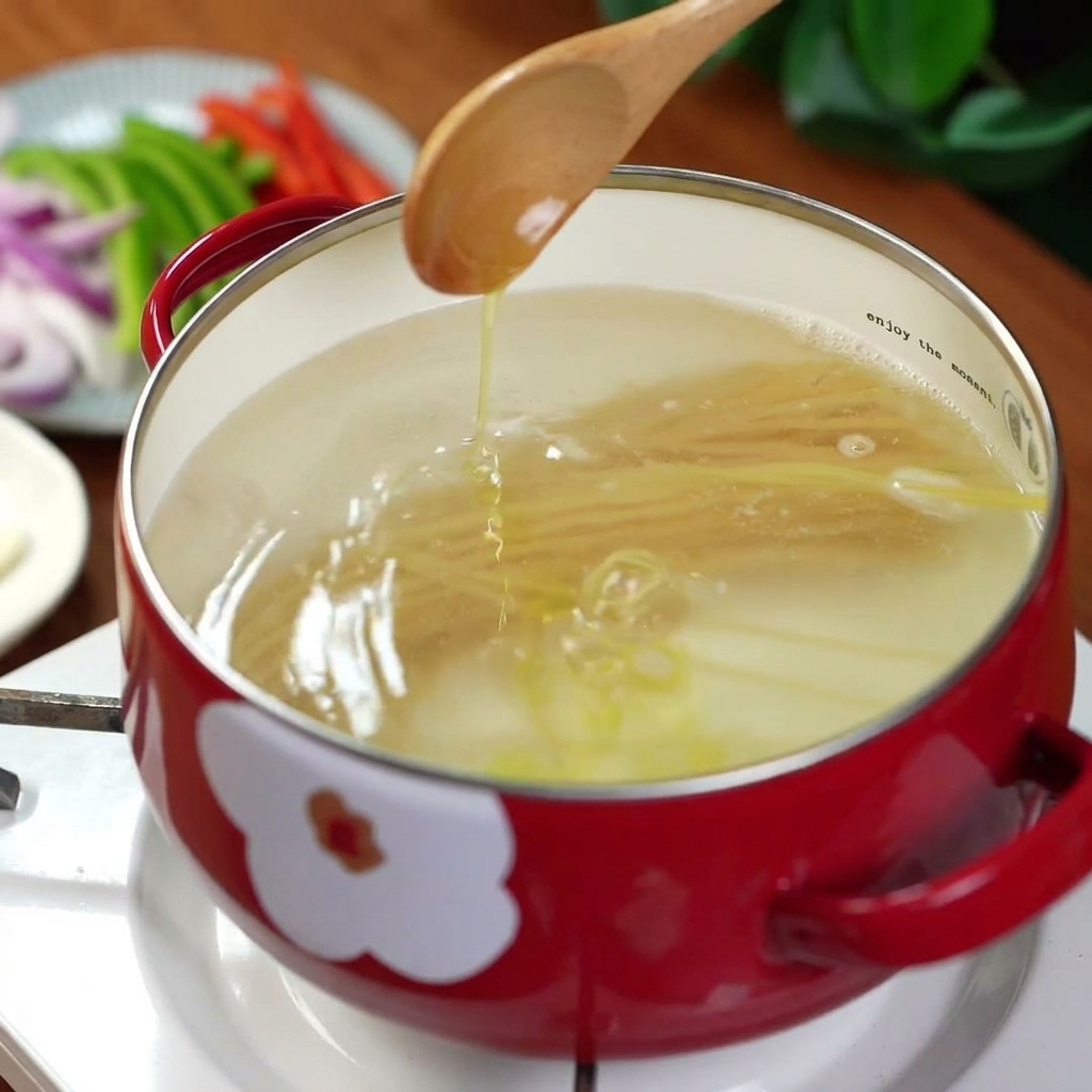 Olio doliva versato da un cucchiaio in una pentola rossa con acqua bollente e spaghetti.