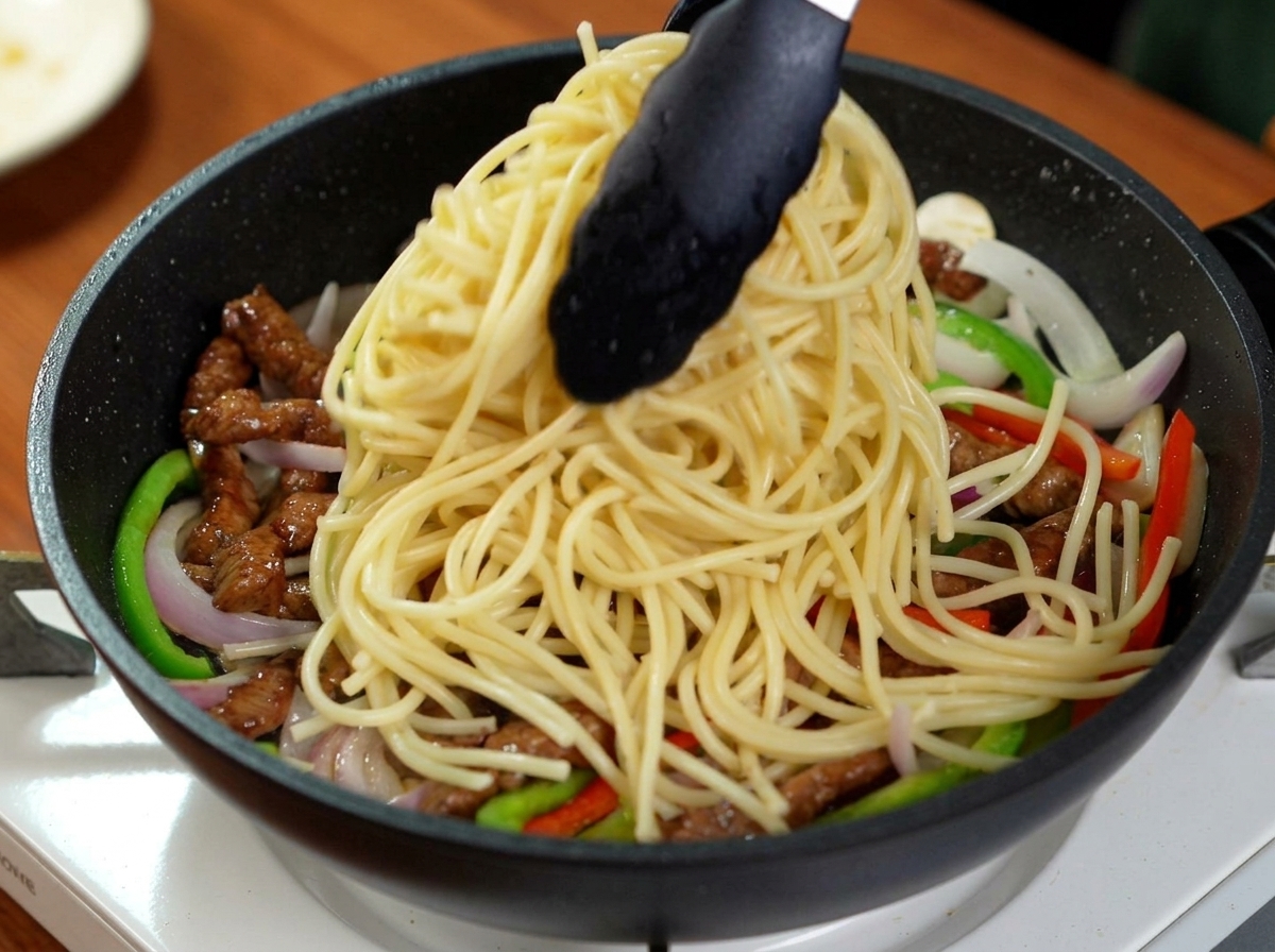Pinze nere che versano spaghetti bolliti in una padella con manzo, peperoni e cipolle.