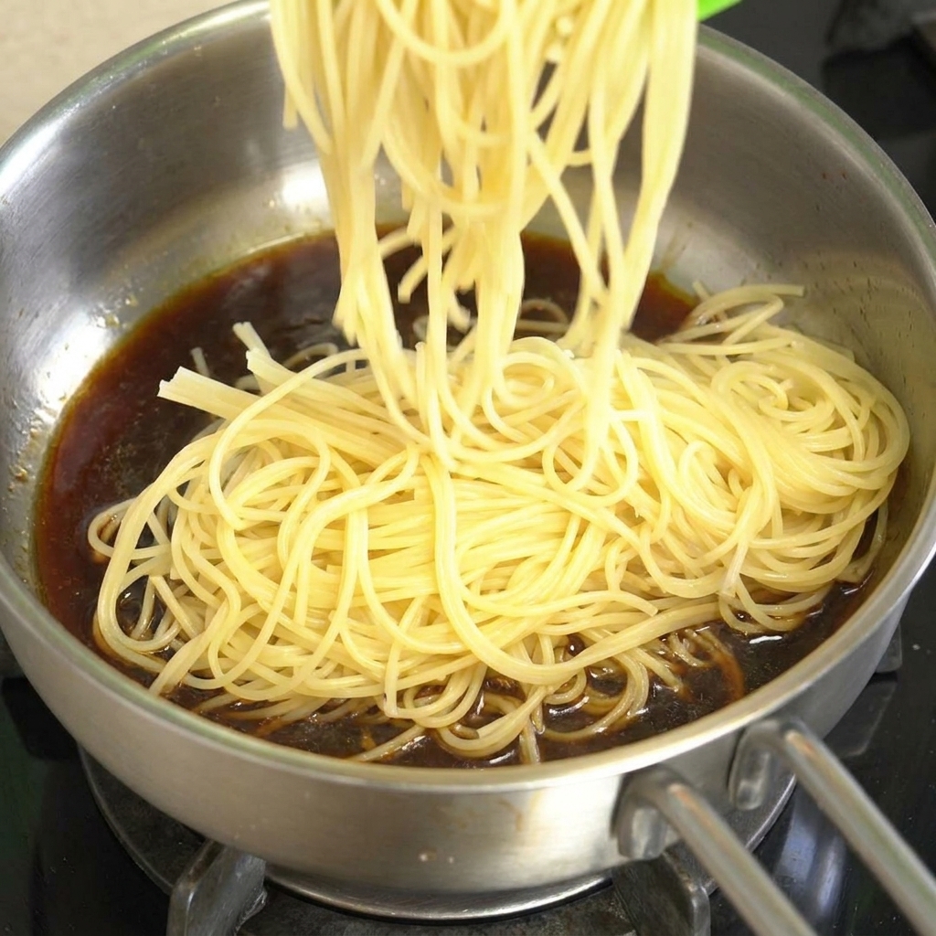Spaghetti cotti che vengono aggiunti in una padella dacciaio contenente salsa scura al pepe nero.