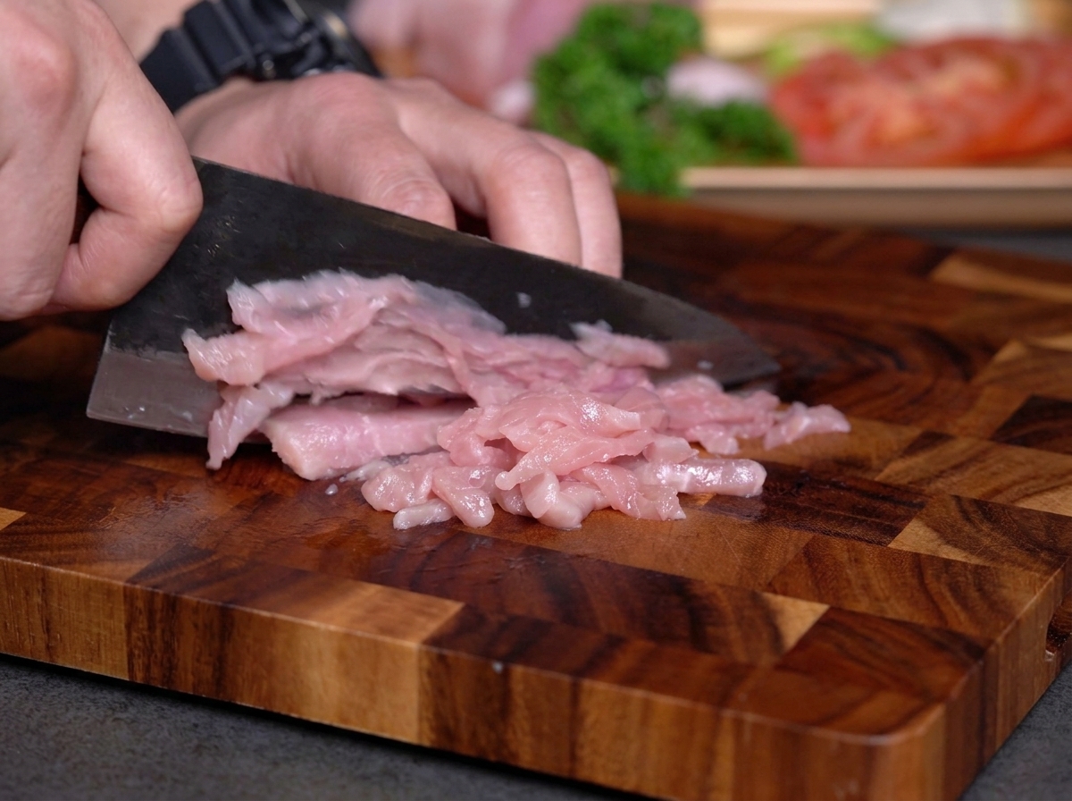 Mani che affettano carne magra cruda a strisce sottili su un tagliere di legno.