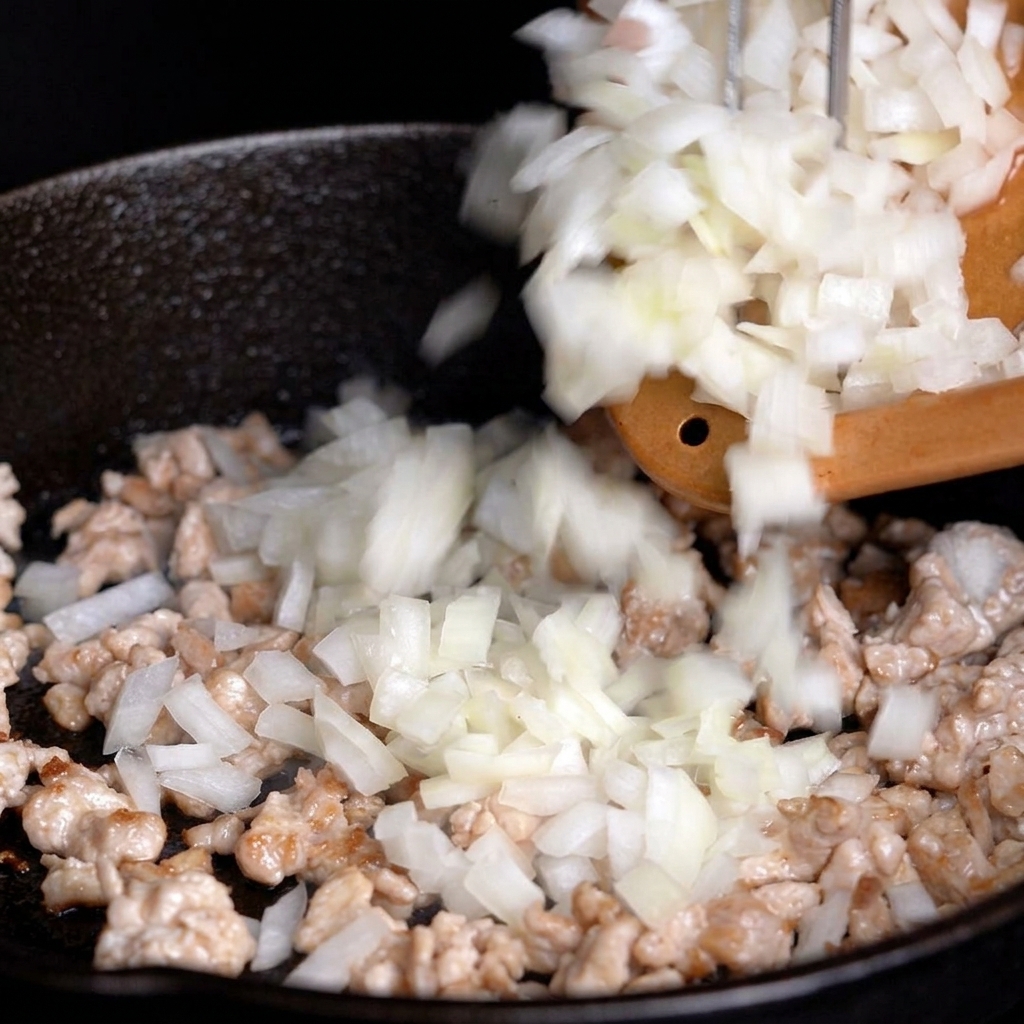 Cipolla bianca tritata versata da un tagliere in una padella con la carne parzialmente cotta.