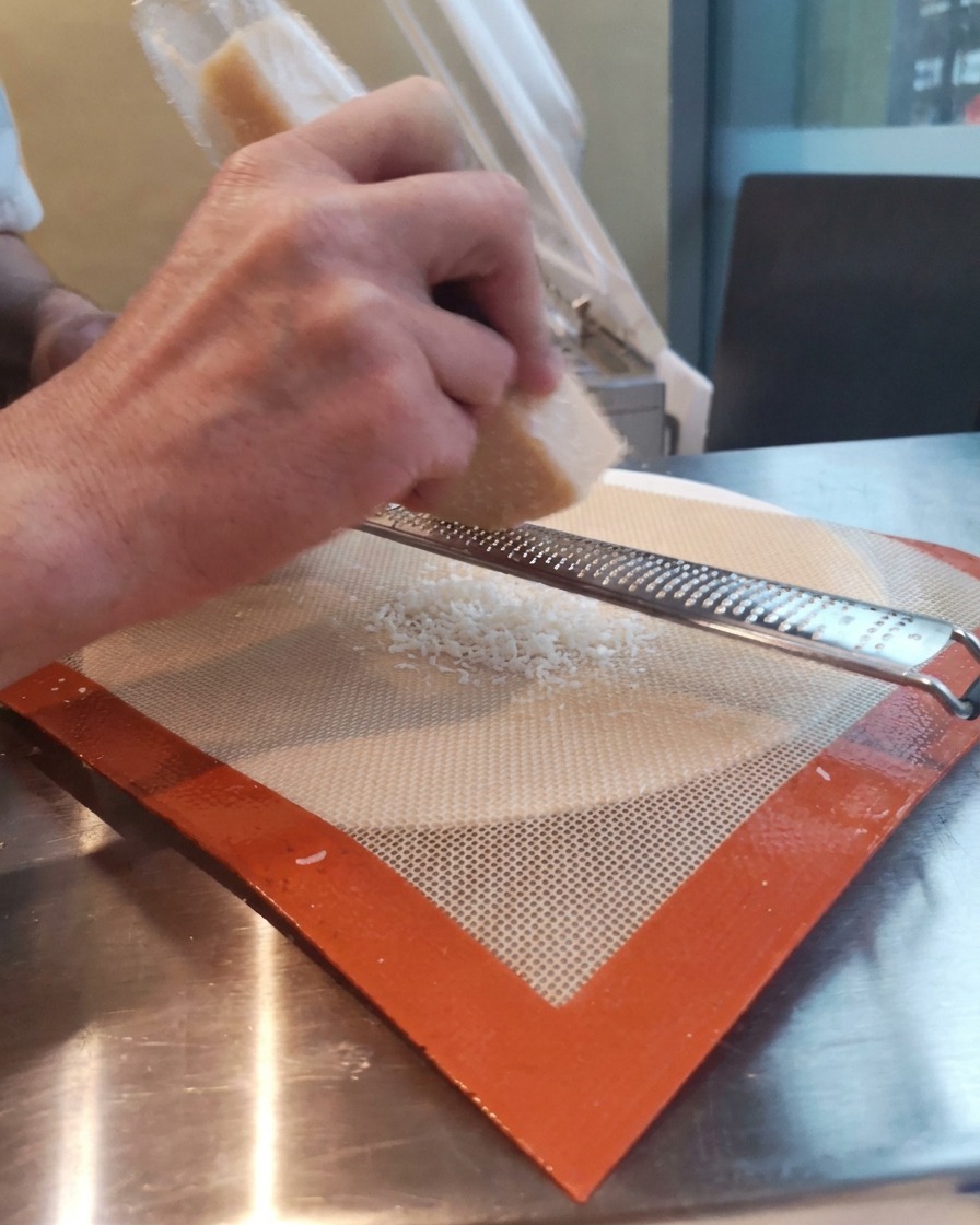 Mani che grattugiano un pezzo di Parmigiano sopra un tappetino in silicone usando una grattugia Microplane.