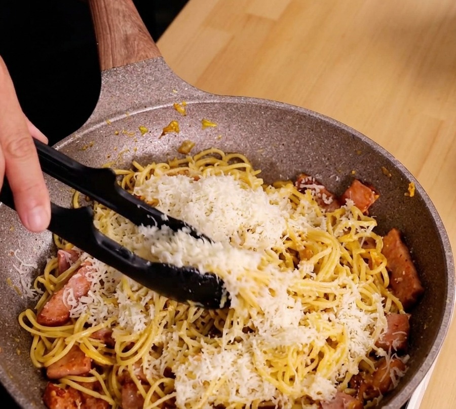 Mani che usano le pinze per saltare energicamente spaghetti, uova e guanciale creando una salsa lucida.
