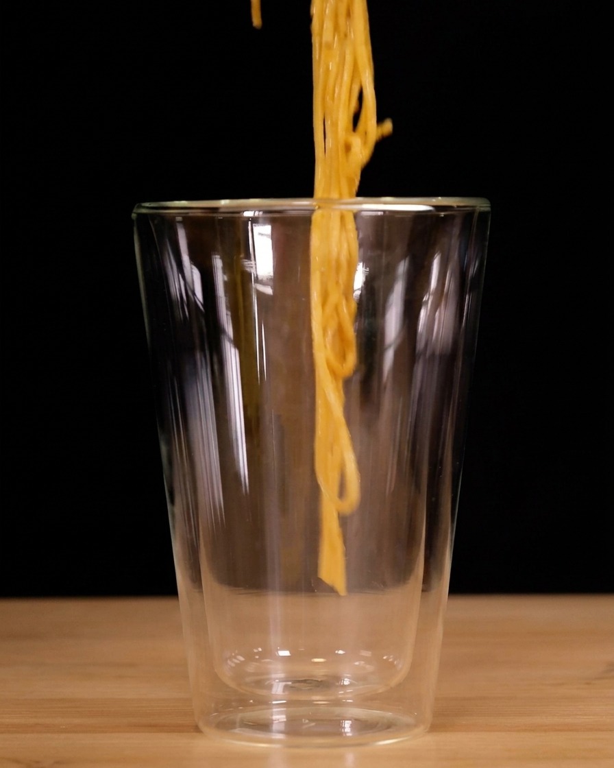 Spaghetti arrotolati verticalmente con le pinze e calati in un alto bicchiere a doppia parete.