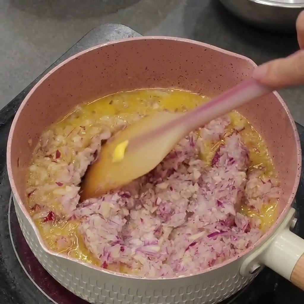 Trito di cipolla rossa e aglio che soffrigge nel burro fuso allinterno di un pentolino rosa.