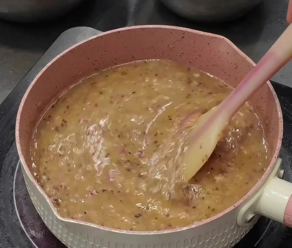 Salsa al pepe nero liquida che bolle in un pentolino rosa, mescolata attivamente con una spatola in silicone.