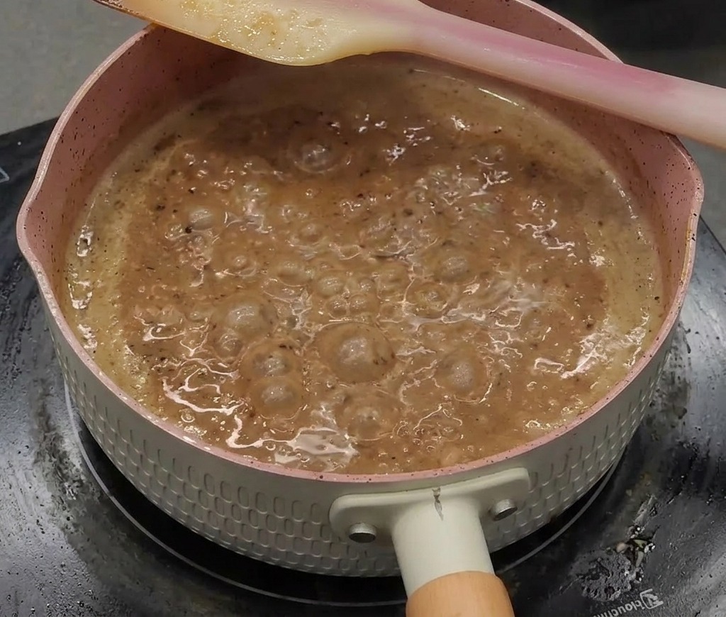 Una salsa al pepe nero densa, lucida e scura che bolle vigorosamente in un pentolino rosa.
