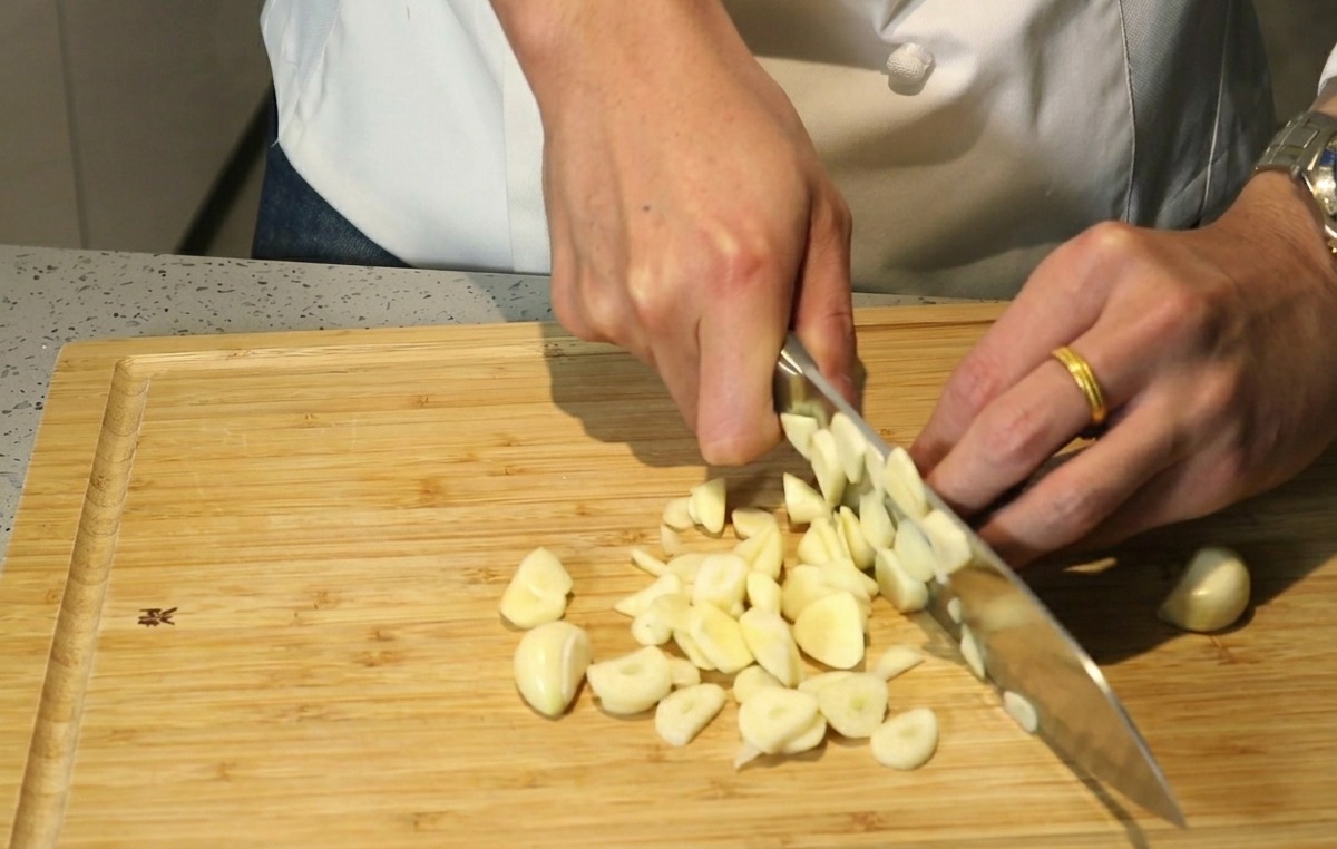 Mani di uno chef che usano un coltello per tritare grossolanamente spicchi daglio su un tagliere di legno.