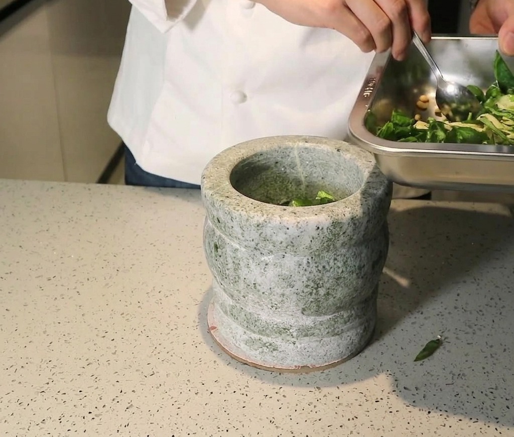 Chef che versa un mix di basilico fresco, pinoli e aglio da un vassoio di metallo in un mortaio di pietra grigia.