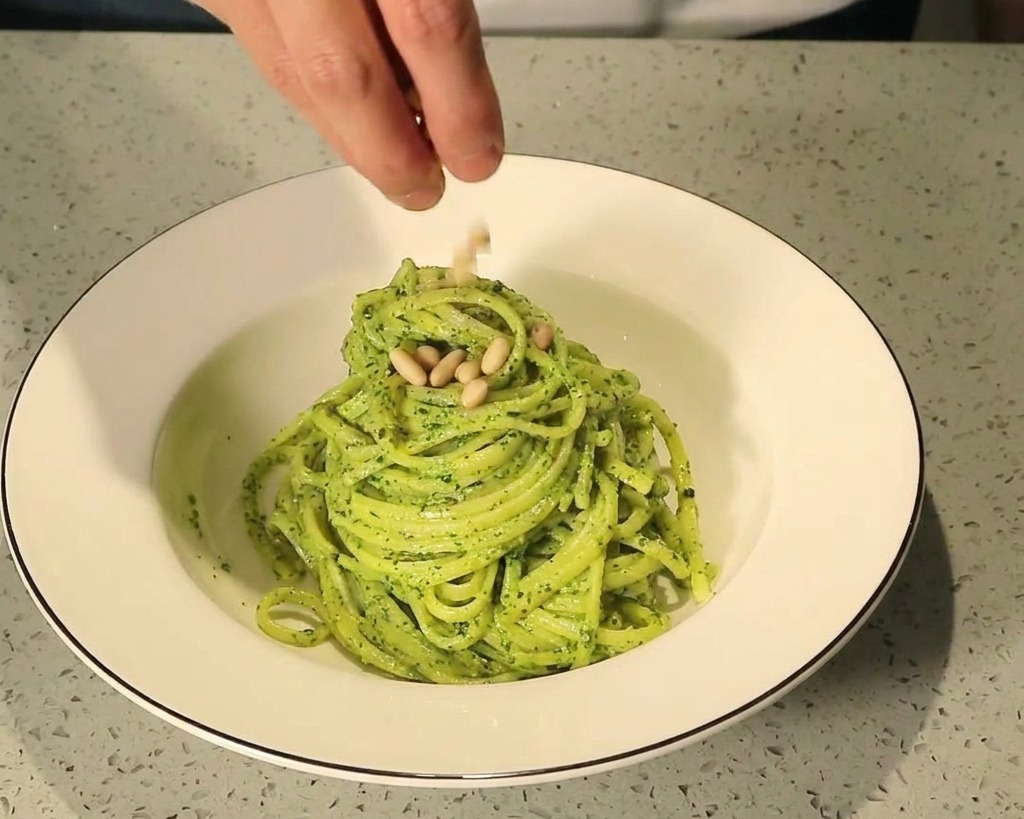 Dita che cospargono pinoli tostati su un nido di pasta al pesto verde in una ciotola di ceramica bianca.