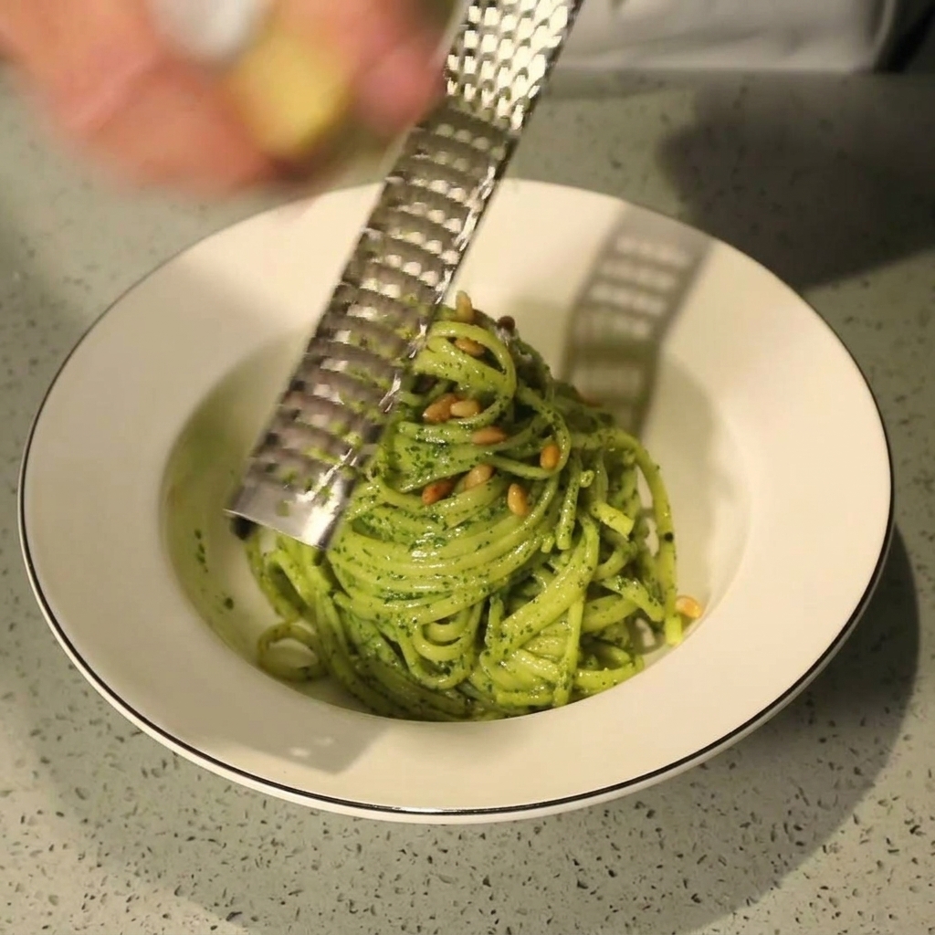 Mano che usa una grattugia per grattugiare Parmigiano fresco su una ciotola di pasta al pesto con pinoli.
